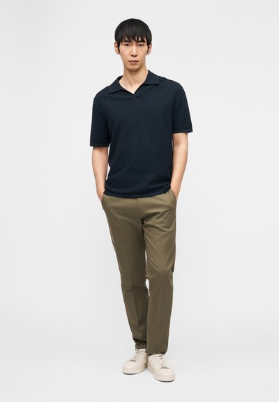 Άνδρας που στέκεται με τα χέρια στις τσέπες, φορώντας μία κοντομάνικη πόλο μπλούζα σε υφή σκούρου ναυτικού μπλε, χακί παντελόνι και λευκά αθλητικά παπούτσια μπροστά σε λευκό φόντο.