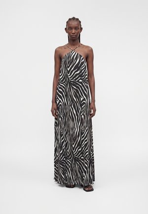 Mujer de pie que lleva un vestido largo de cuello halter con rayas de cebra en blanco y negro y sandalias negras sobre un fondo liso.