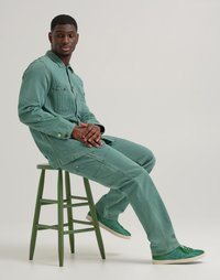 Green corduroy workwear outfit with a button-up jacket and matching pants. Worn with green sneakers, seated on a green wooden stool.