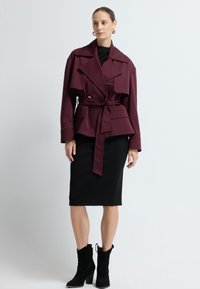 Bordeaux farvet jakke med bælte, store klaplommer og slidser, båret over en tætsiddende sort kjole. Sorte ankelstøvler fuldender outfittet.