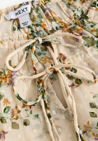 Tissu crème à motif floral, avec des fleurs vertes et orange, et deux cordons de tirage noués sous une taille froncée avec une étiquette de marque NEXT.
