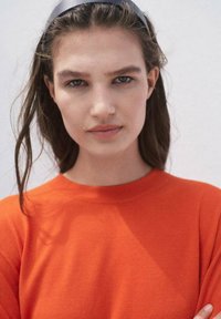 Helle orange, kurzärmlige Strickoberteil mit Rundhalsausschnitt. Das Modell hat welliges braunes Haar, das mit einem dunklen Haarband gestylt ist.