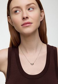 Srebrny naszyjnik z wisiorkiem w kształcie serca na cienkim łańcuszku, noszony na ciemno brązowej bluzce bez rękawów. Gładka powierzchnia, minimalistyczny design.