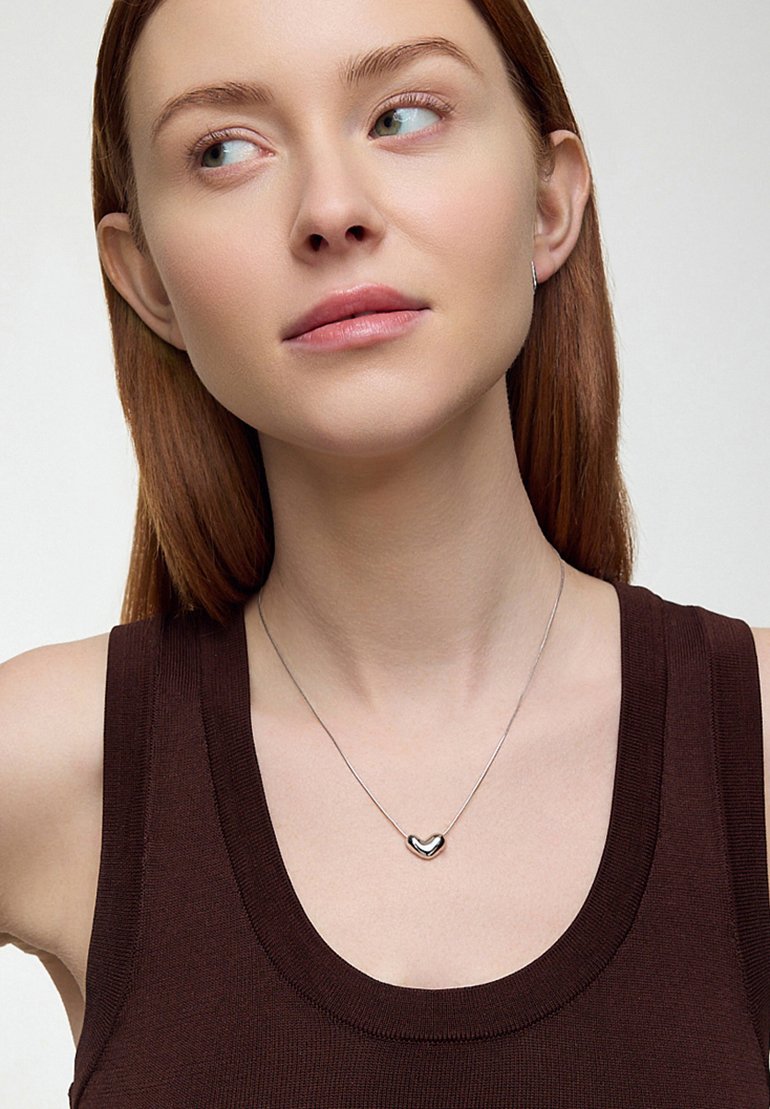 Srebrny naszyjnik z wisiorkiem w kształcie serca na cienkim łańcuszku, noszony na ciemno brązowej bluzce bez rękawów. Gładka powierzchnia, minimalistyczny design.
