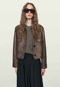 Žena s vlnitými vlasy, oblečená v nadměrných tmavých slunečních brýlích, hnědé kožené bundě přes šedý svetr a černé plisované sukni, neutrální pozadí.