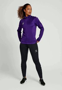 Vijolična športna jopica z zadrgo in stranskimi žepi, kombinirana s črnimi oprijetimi legicami. Superge imajo črno-bele detajle, ki zagotavljajo športen videz.