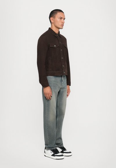 Veste en daim marron avec fermeture à boutons et deux poches poitrine, associée à un jean bleu délavé et des baskets noir et blanc.