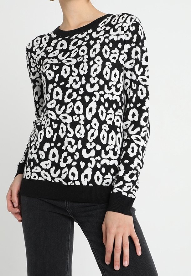Femme portant un pull à manches longues imprimé léopard noir et blanc et un pantalon noir, avec une main posée sur sa cuisse.