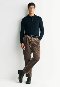 Black long-sleeve polo shirt, tan corduroy pants with visible pleats, black belt, and black shoes with thick soles.