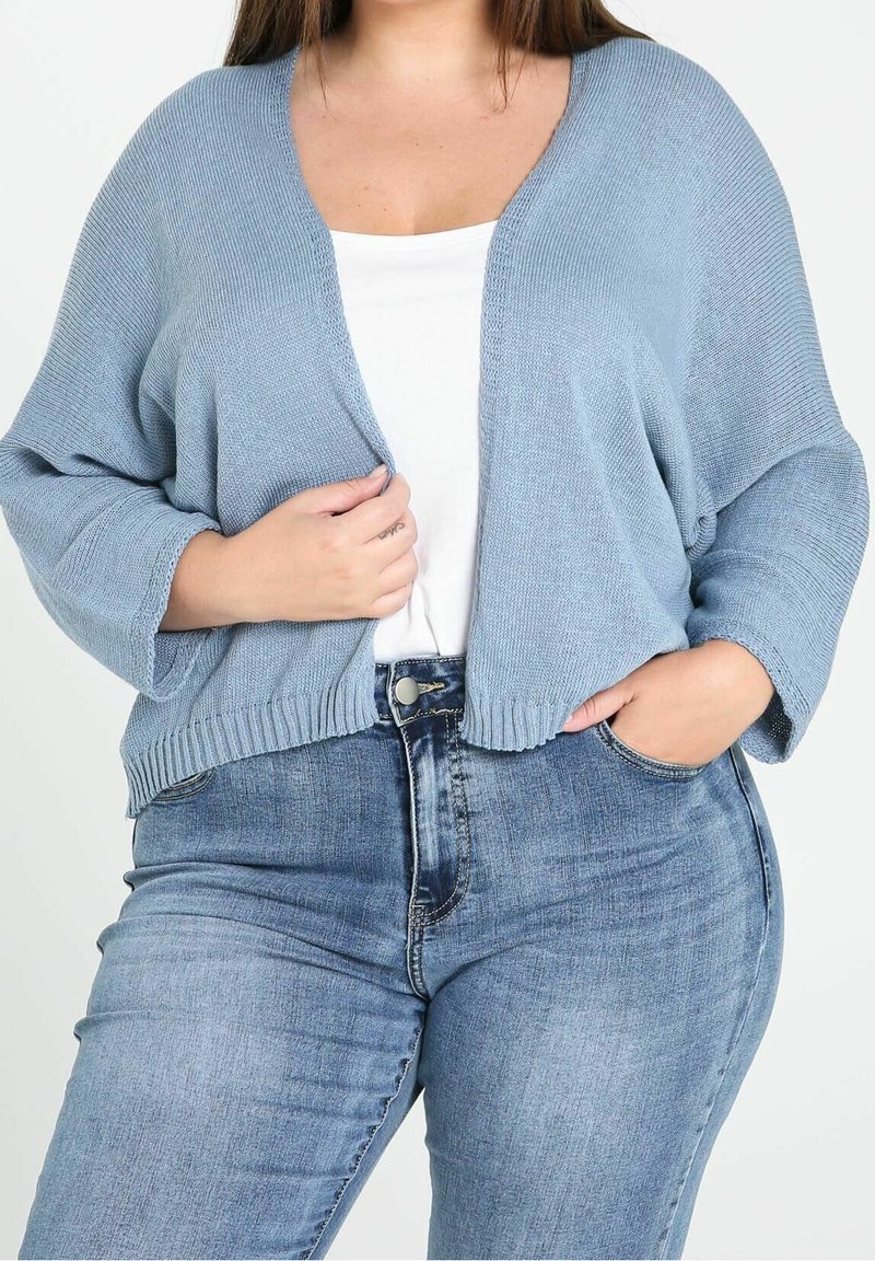 Jasnoniebieski sweter na drutach z dekoltem w serek i szerokimi rękawami, w połączeniu z białym t-shirtem i dopasowanymi niebieskimi dżinsami. Dzianina o teksturowanym wzorze.