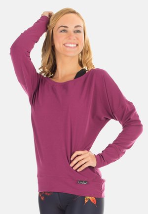 Lachende vrouw met blond haar draagt een losse paarse top met lange mouwen en donkere legging met oranje bladpatronen, poseert met één hand op haar heup.