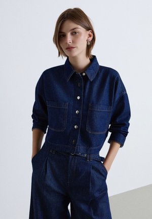 Jeune femme aux cheveux courts portant une veste en jean bleu foncé et un jean taille haute assorti, debout avec les mains dans les poches devant un fond uni.