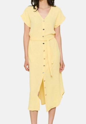 Kvinde iført en lysegul skjortekjole med korte ærmer, V-hals og bælte bundet i taljen, stående foran en ensfarvet baggrund.