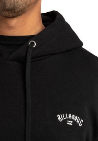 Sudadera negra de tela suave. Cuenta con un bolsillo frontal y capucha con cordón, con el logo bordado en blanco "BILLABONG" en el pecho.
