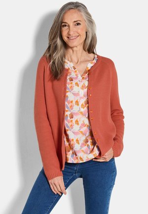 Cardigan en maille corail avec un design texturé et des boutons orange, assorti à une chemise à imprimé floral et un jean bleu.