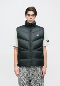 Mustast pehmendusega vest, millel on kõrge krae, lukk ja küljetaskud. Kandmiseks beezist särgi ja mustriliste heledate pükstega. Nike logo on nähtaval.