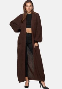 Lång, brun stickad cardigan med lös passform, vida ärmar, två framfickor och ett texturerat mönster. Bärs över en svart crop top och byxor.