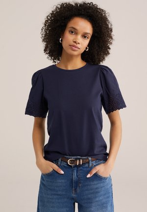 Kobieta o kręconych włosach, ubrana w granatową bluzkę z bufkami i ażurowymi zdobieniami, niebieskie jeansy oraz brązowy pasek, stojąca z rękami w kieszeniach.