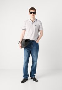 Polo cinza claro com detalhes em preto, jeans azuis e sapatos pretos. O modelo segura uma bolsa de couro preta e usa óculos de sol escuros.