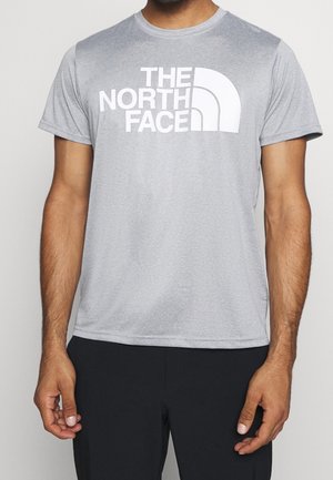 Mężczyzna w jasnoszarej koszulce z krótkim rękawem z dużym białym logo "THE NORTH FACE" i czarnych spodniach, stojący na jednolitym tle.