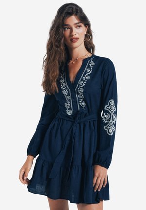 Marineblaues Kleid aus weichem Stoff, mit weißer Blumenstickerei, einer durchgängigen Knopfleiste, langen Ärmeln und einem taillierten Bund, der in einen gestuften Rock übergeht.