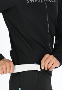 Svart zip-up-jacka med ett vitt elastiskt band i midjan, med en logotyp på bröstet och blå detaljer på sidan.