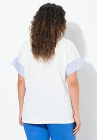 Valkoinen T-paita, jossa on lyhyet sinivalkoiset raidoitetut hihat, rento leikkaus ja pehmeä materiaali, yhdistettynä yksinkertaisiin sinisiin housuihin.