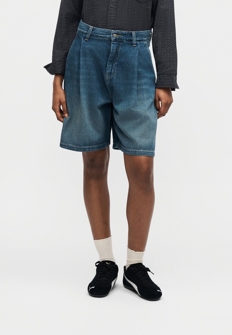 Persona che indossa pantaloncini di jeans blu sbiaditi di lunghezza media, maglietta nera a maniche lunghe, calzini bianchi e scarpe da ginnastica nere, in piedi su sfondo uniforme.