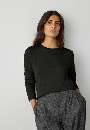 Schwarzes Langarmoberteil mit gerippter Textur und Rundhals-Ausschnitt, kombiniert mit hochgeschnittenen, grauen karierter Hose, Hände in den Taschen.