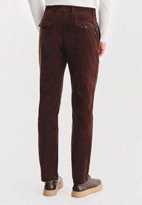 Brązowe zamszowe spodnie o slim fit, z dwoma tylnymi kieszeniami i subtelnymi detalami w szwie. W zestawieniu z ciemnobrązowymi butami.