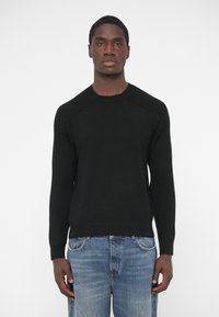 Čierny pletený sveter s rebrovanými detailmi na ramenách a manžetách, s okrúhlym krkom, v kombinácii s modrými džínami, nosený mužským modelom.