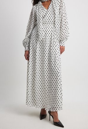Mujer con un vestido largo blanco con lunares negros, mangas largas, una lazada tipo pañuelo en el cuello y tacones altos negros y puntiagudos.