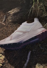 Scarpa da ginnastica rosa chiaro con suola testurizzata appoggiata su un terreno boschivo coperto di terra, foglie e piccoli rami vicino all'erba verde.