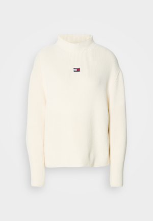 Cream turtleneck sweater with ribbed texture and a small colored logo patch on the chest. Features loose-fitting sleeves and a relaxed shape.