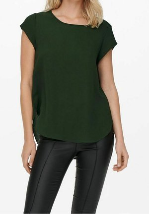 Femme portant un chemisier vert foncé à manches courtes et un pantalon noir ajusté, debout les bras relâchés le long du corps.