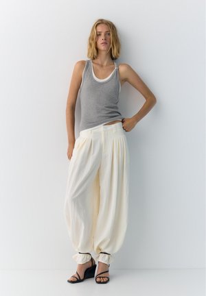 Femme aux cheveux blonds mi-longs porte un débardeur gris sur un sous-t-shirt blanc, un pantalon blanc ample serré aux chevilles, des sandales à talons compensés noires, debout.