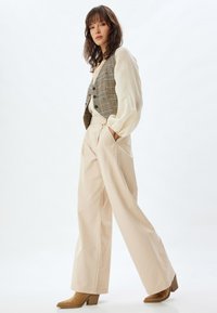 Beige wijde pijpen broek gecombineerd met een geruiten vest; het vest heeft donkere knopen en eronder een lichte crèmekleurige blouse. Bruine hoge laarzen.