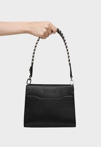 Czarna torebka o fakturze z plecionym czarno-białym uchwytem. Posiada wytłoczone logo "TOUS" z przodu oraz usztywnioną prostokątną formę.