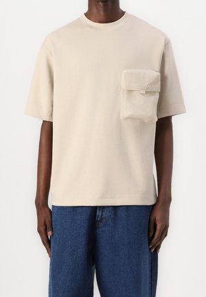 Mørkhudet person iført en beige kortærmet T-shirt med en stor forreste lomme og blå cowboybukser mod en ensfarvet baggrund.