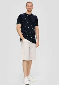 Marine t-shirt met groen sterrenpatroon, gecombineerd met lichtbeige shorts. Model draagt witte sneakers en staat met de handen in de zakken.