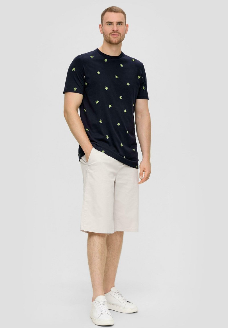 Marine t-shirt met groen sterrenpatroon, gecombineerd met lichtbeige shorts. Model draagt witte sneakers en staat met de handen in de zakken.