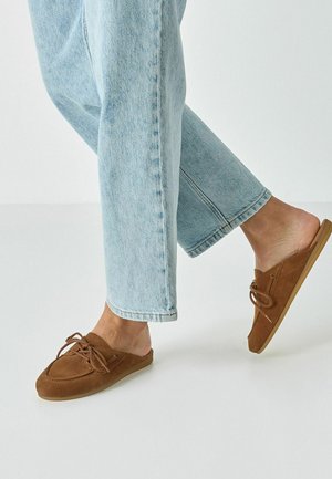 Bruine suède instapschoenen met veters, ronde neus en rubberen zool, gecombineerd met lichtblauwe jeans in een minimalistische omgeving.