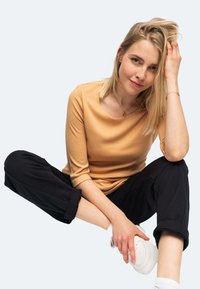 Beige Langarmshirt mit Rundhalsausschnitt, kombiniert mit schwarzen Hosen mit umgeschlagenen Säumen. Weiße Sneakers mit durchbrochenen Details vervollständigen das Outfit.