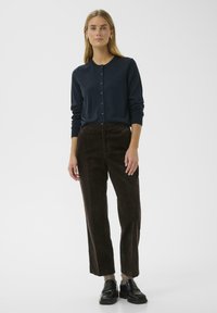 Cardigan marine avec boutons à l'avant, associé à un pantalon en velours côtelé marron à coupe droite. Le mannequin porte des mocassins noirs, mettant en valeur un tissu texturé et une coupe décontractée.