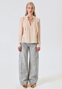 Blusa beige con dettagli arricciati, scollatura a V profonda e maniche lunghe; abbinata a jeans grigi chiari a gamba larga e scarpe con dettaglio a fiocco dorato.