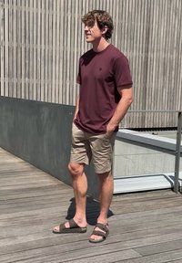 Jeune homme aux cheveux bouclés debout à l’extérieur portant un t-shirt bordeaux, un short beige et des sandales marron, les mains dans les poches.