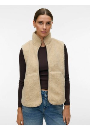 Donna che indossa un gilet in pile beige con cerniera sopra una maglia nera a maniche lunghe e jeans blu, in piedi davanti a uno sfondo bianco semplice.