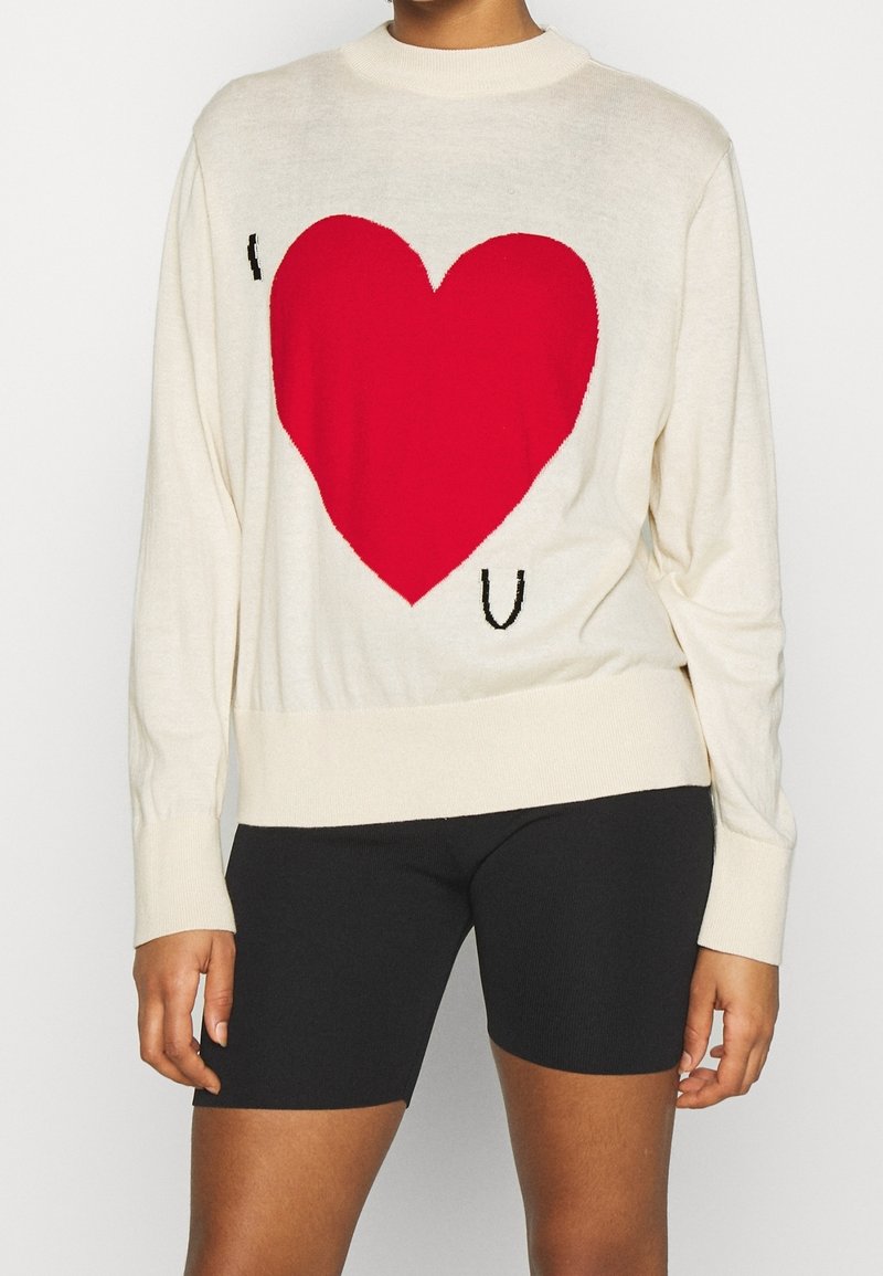 Femme portant un pull blanc avec un grand cœur rouge et les lettres "I" et "U" à l'avant, associé à un short de cycliste noir.