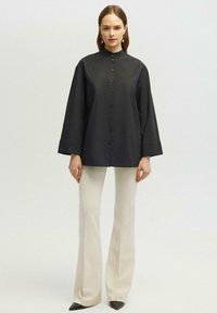 Camisa negra de botones con un alto cuello y mangas amplias, combinada con pantalones anchos crema y zapatos negros de punta. Textura de tela suave.