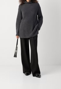 Šedý žebrovaný svetřík s vysokým límcem, doplněný černými kalhotami s širokými nohavicemi. Doplňky zahrnují průhlednou kabelku a černé baleríny s cvočky.